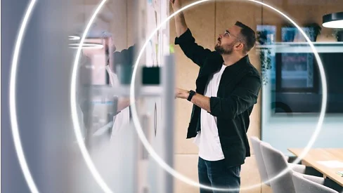 A man stands and writes on a whiteboard in a modern office setting. There are concentric circular light effects over the image. He is wearing a black jacket and white t-shirt. A conference table and chairs are visible in the background, with some tesa tape neatly placed near the edge for office use. (Acest text a fost generat de inteligența artificială)