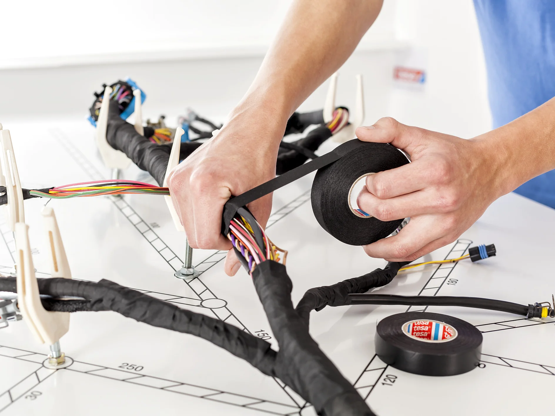 Hands are wrapping black tesa tape around a bundle of colorful electrical wires. The workspace is marked with measurements, and a small roll of tesa tape and various connectors are visible. (Acest text a fost generat de inteligența artificială)