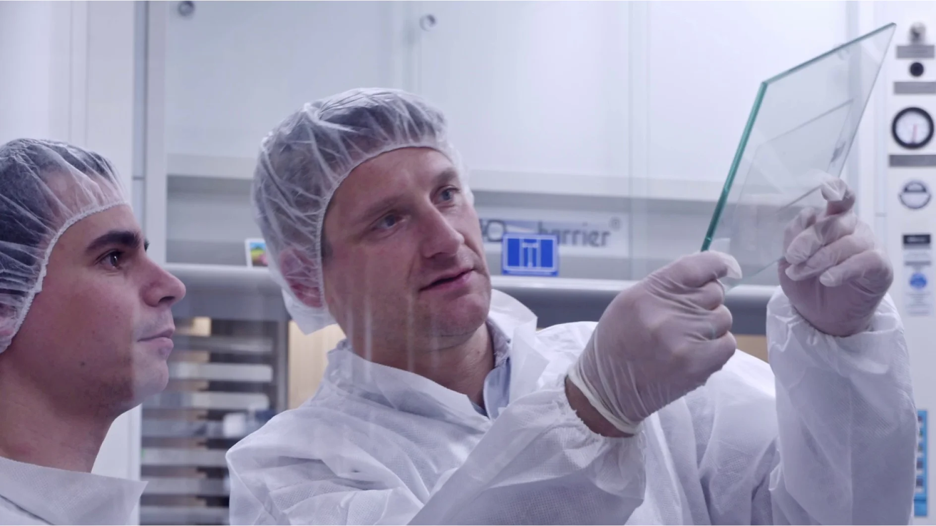 Two people in protective lab clothing and hairnets, one holding and examining a transparent sheet of tesa tape. They appear to be in a laboratory setting with equipment visible in the background. (Acest text a fost generat de inteligența artificială)