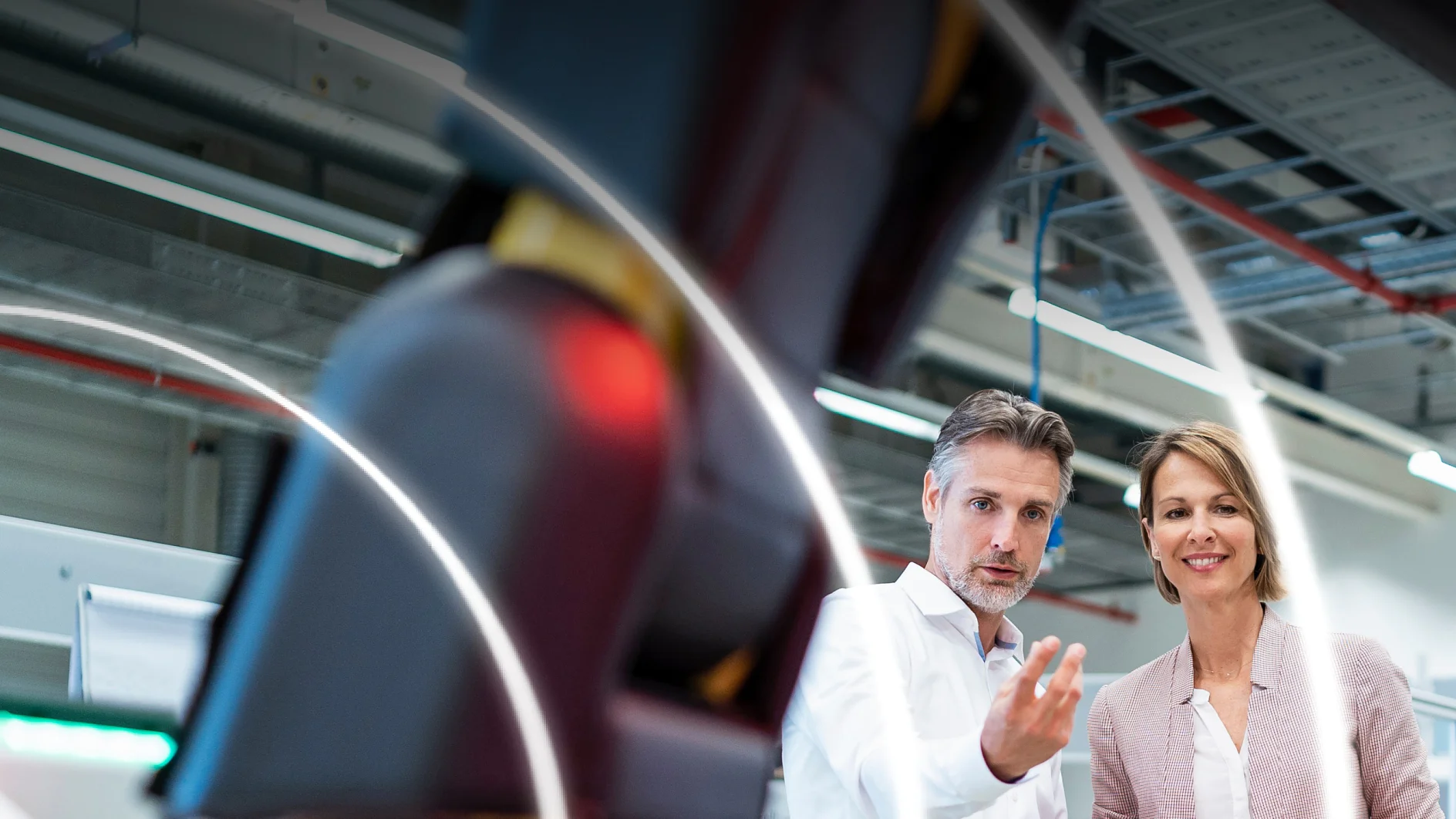 Two people in a modern industrial setting observe a large robotic arm in the foreground. The man gestures with his hand, while the woman stands beside him, both appearing engaged with the machinery. Visible are overhead cables and lighting secured with tesa tape. (Acest text a fost generat de inteligența artificială)