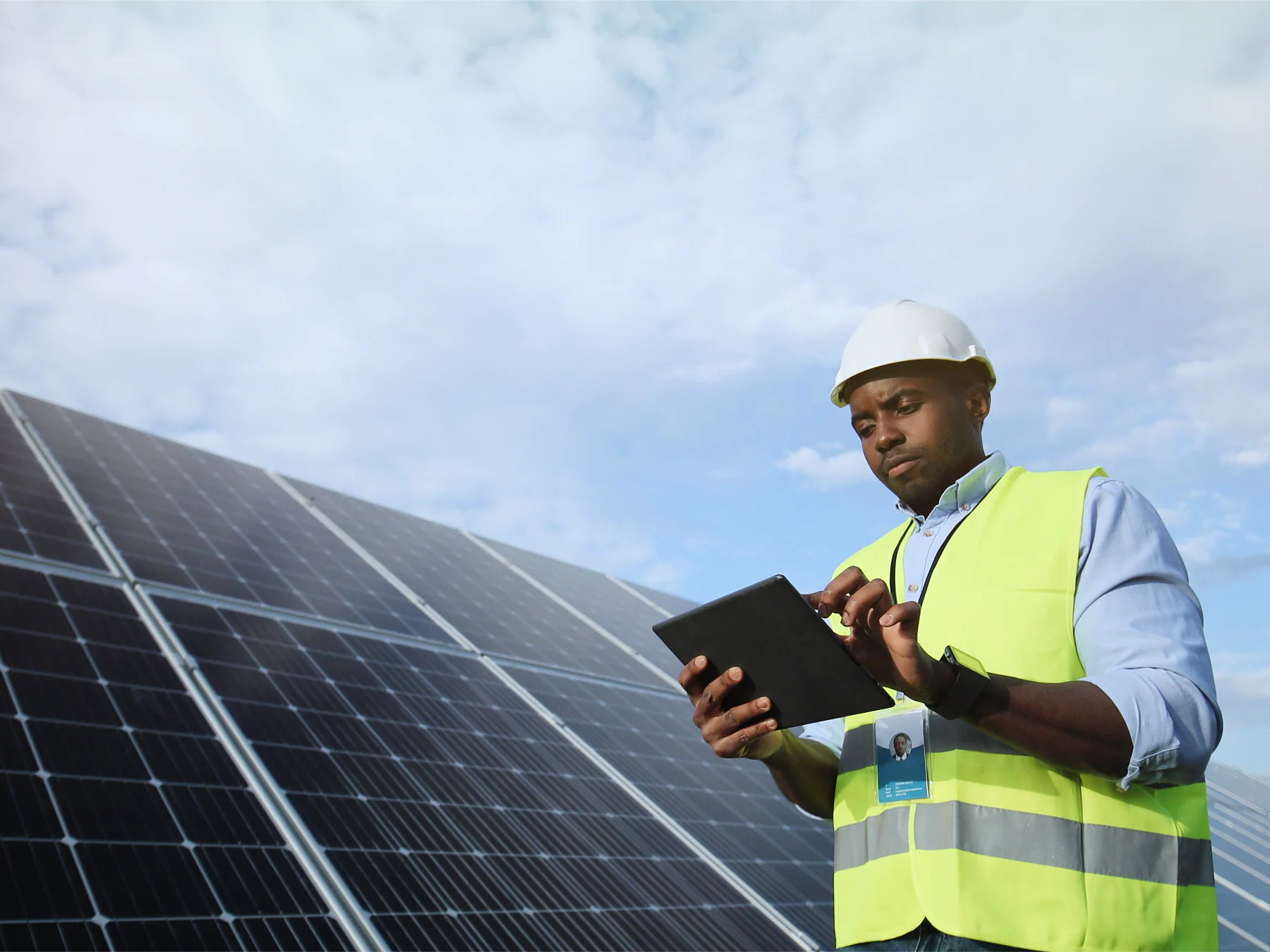 A person wearing a hard hat and neon safety vest is using a tablet near large solar panels secured with tesa tape. The sky is partly cloudy. (Acest text a fost generat de inteligența artificială)