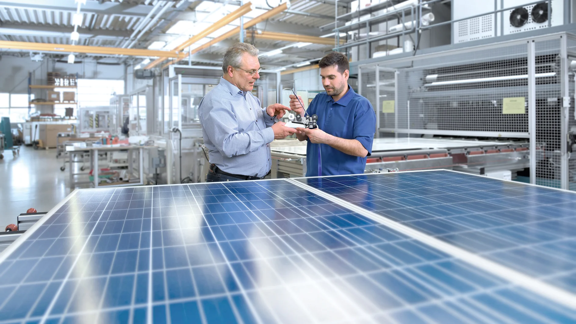 Two men are examining a small piece of machinery in a bright, industrial setting. They stand next to large solar panels. The background shows various equipment and metal structures, indicating a technological or manufacturing environment. (Acest text a fost generat de inteligența artificială)