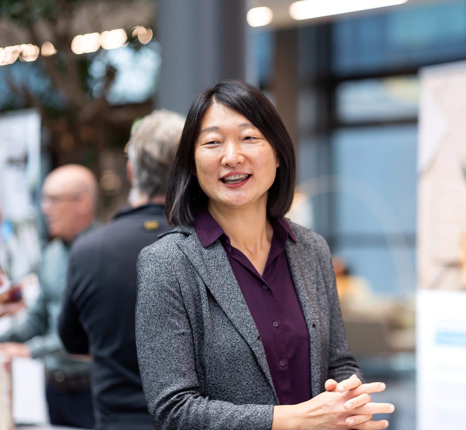 A woman with shoulder-length black hair is smiling, wearing a gray blazer and purple blouse. She is standing indoors, with people and blurred background elements like posters and a window behind her. Please provide the text that contains references to tapes so I can help you make the necessary modifications by using tesa tape. (This text has been generated by AI)