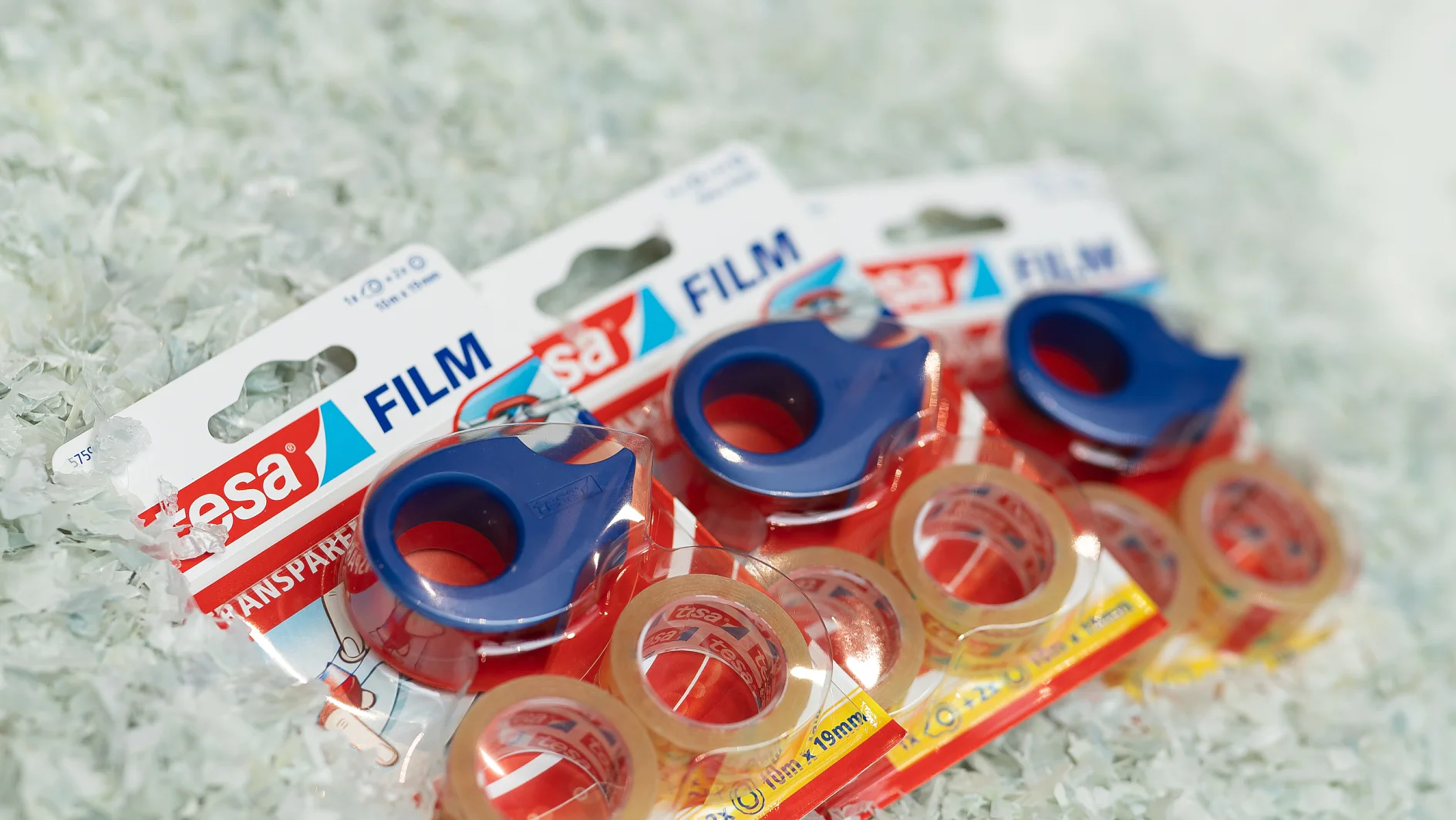 Three packages of transparent tesa tape with blue dispensers are displayed. Each package includes three rolls of tesa tape and features brand labeling. The background is covered with shredded white material. (This text has been generated by AI)