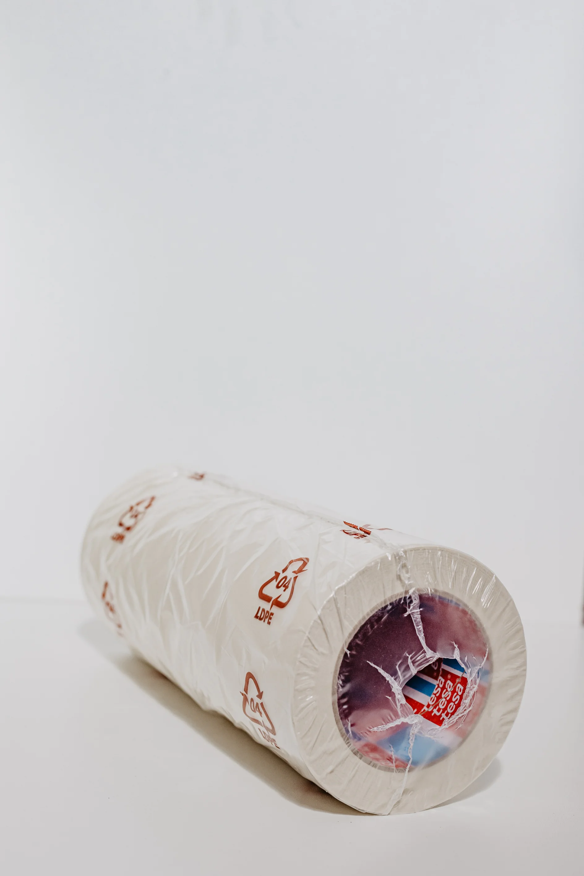 A roll of white plastic wrap is standing upright against a plain white background. The packaging is partially transparent, revealing the inner cardboard tube and multicolored label. Red recycling symbols and text can be seen on the plastic. (This text has been generated by AI)