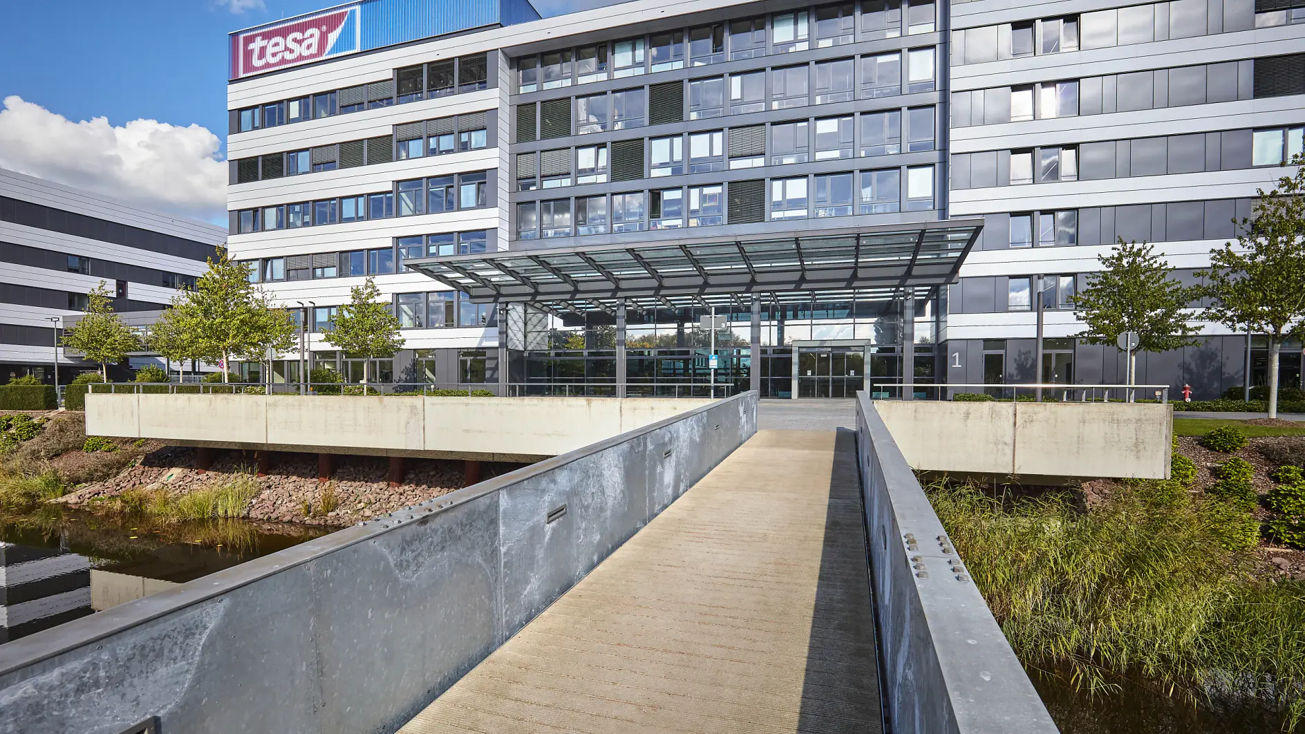 Exterior view of a modern office building with a glass and metal facade. A pedestrian bridge leads to the main entrance, featuring a glass canopy. The building is surrounded by greenery and water, and a tesa tape logo is visible at the top. (Acest text a fost generat de inteligența artificială)