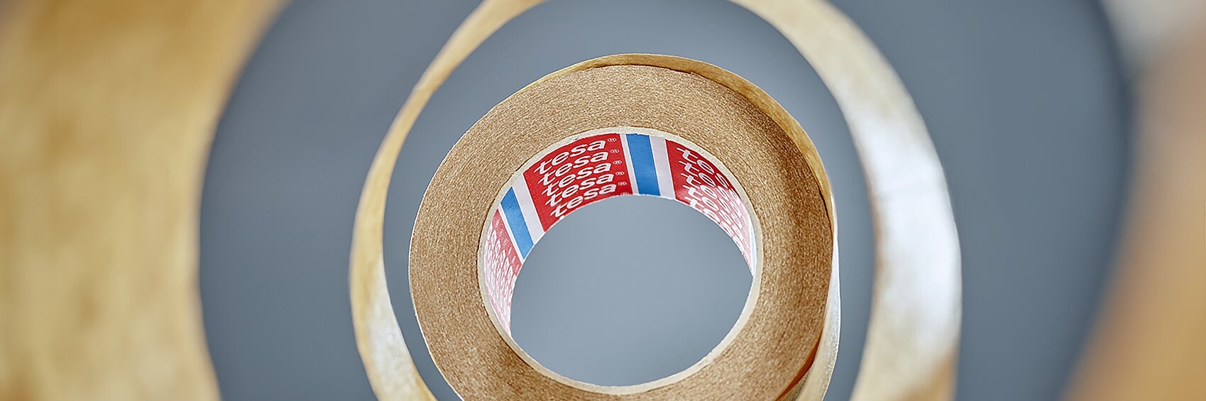 View from the center of a roll of tesa tape, creating a spiral pattern. The tesa tape is brown with a partially visible red label inside, featuring text and colored stripes. The background is blurred, emphasizing the layered effect of the tesa tape. (This text has been generated by AI)