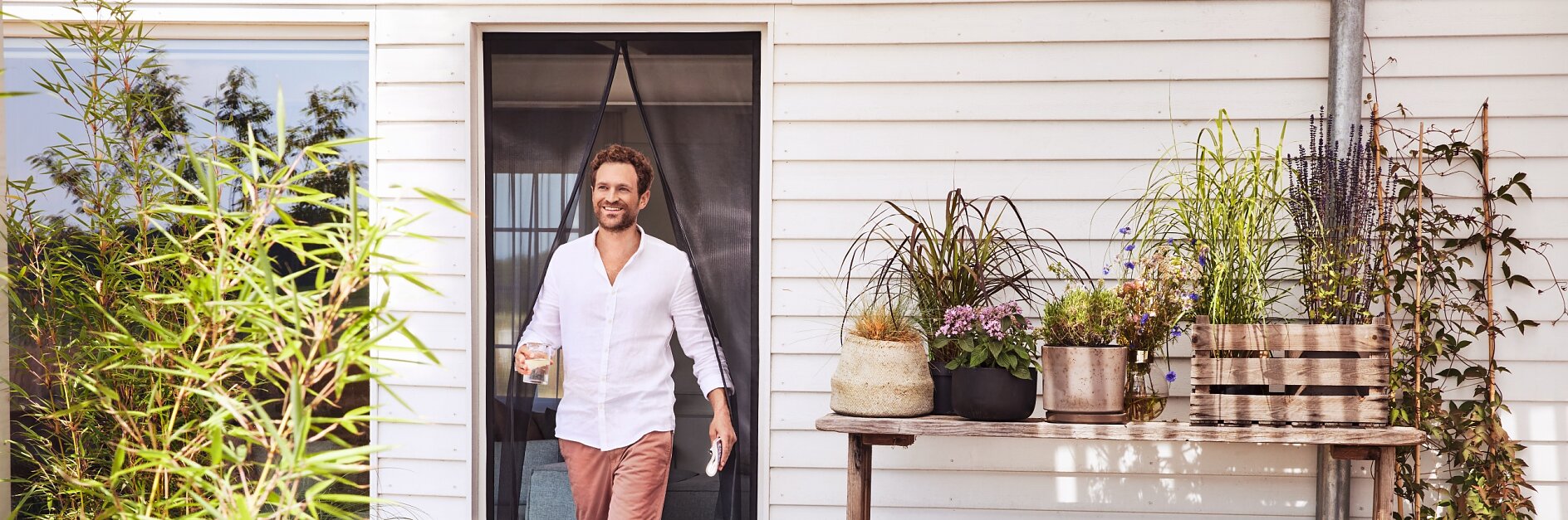 A man wearing a white shirt and pink pants walks through a door onto a wooden deck, holding a glass. The deck features potted plants, a stack of firewood, and a table with more plants. The building has white siding. (This text has been generated by AI)