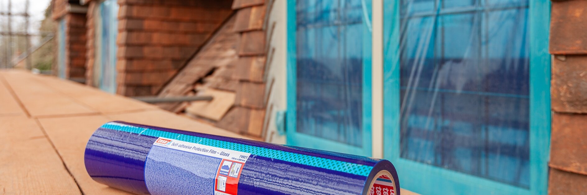 O rolă de membrană adezivă tesa tape albastră se află pe o suprafață de lemn în exteriorul unei clădiri în construcție, cu ferestre cu rame albastre și siding maro vizibile în fundal. (Acest text a fost generat de inteligența artificială)