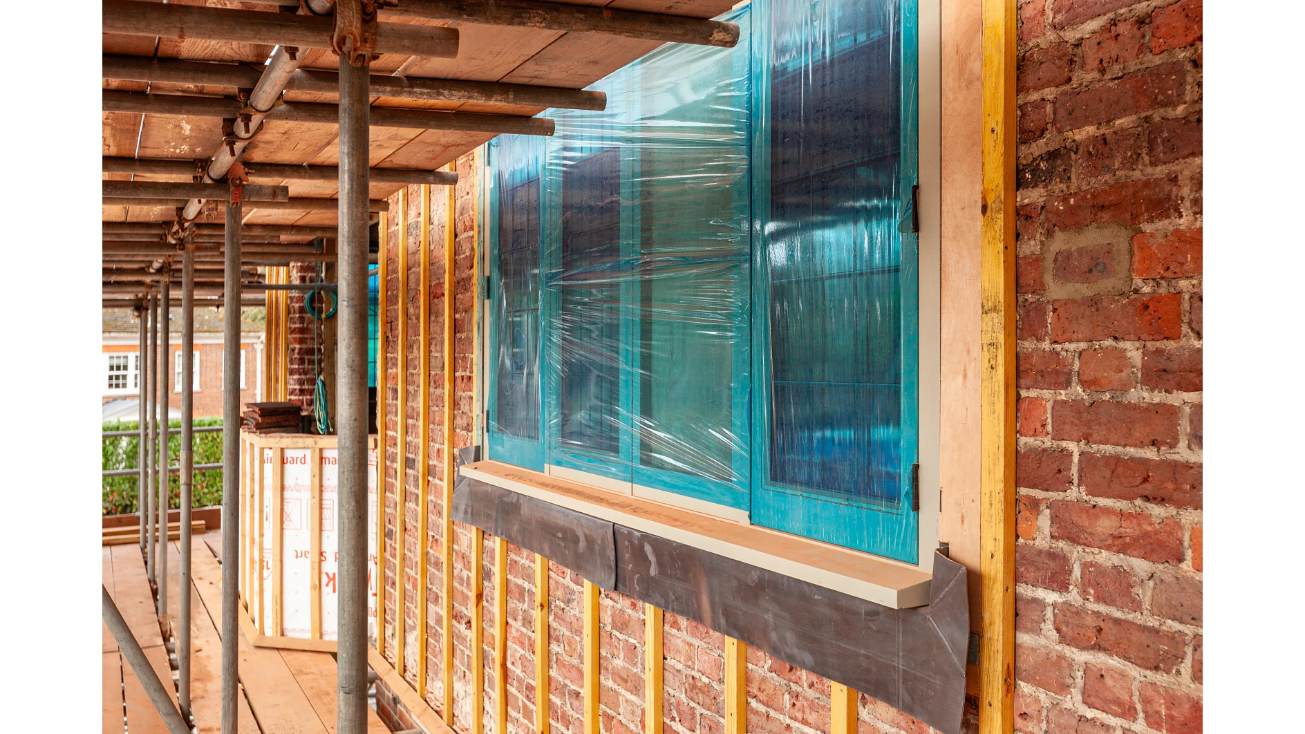 Clădire din cărămidă în construcție sau renovare, cu schele, structură din lemn expusă și o fereastră acoperită cu plastic de protecție albastru fixat cu bandă tesa. Unele materiale de construcție sunt vizibile în fundal. (Acest text a fost generat de inteligența artificială)