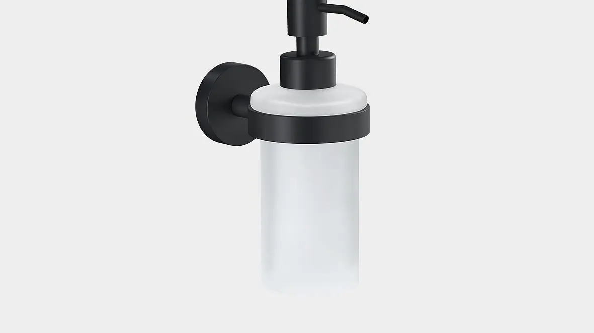 Distribuitor de săpun montat pe perete, cu un recipient din sticlă mată și pompă și suport din metal negru. Dozatorul este fixat cu bandă tesa pe un fundal alb simplu.