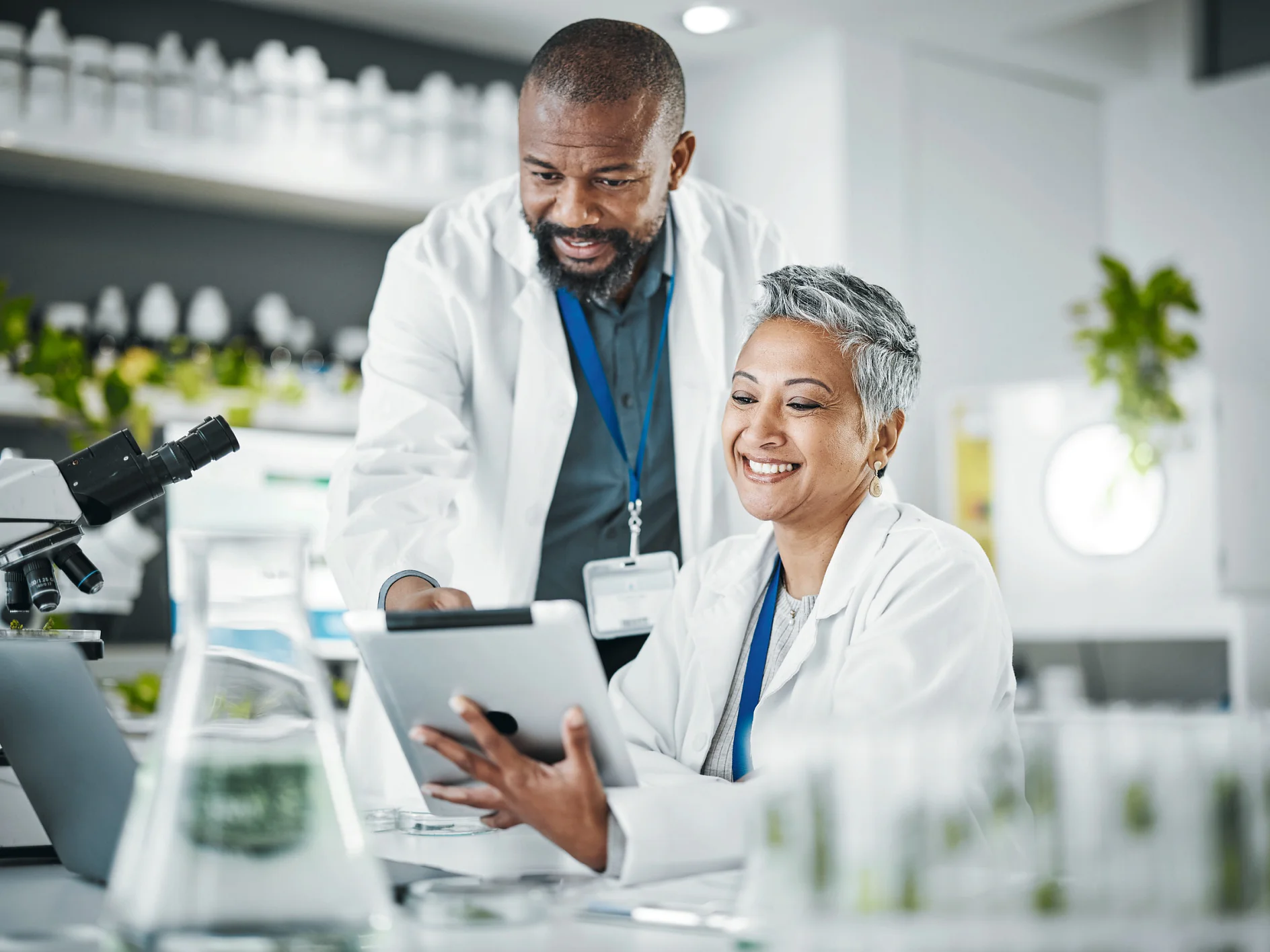 Two scientists in white lab coats using a tablet in a bright laboratory with microscopes and glassware on the table tesa (Acest text a fost generat de inteligența artificială)