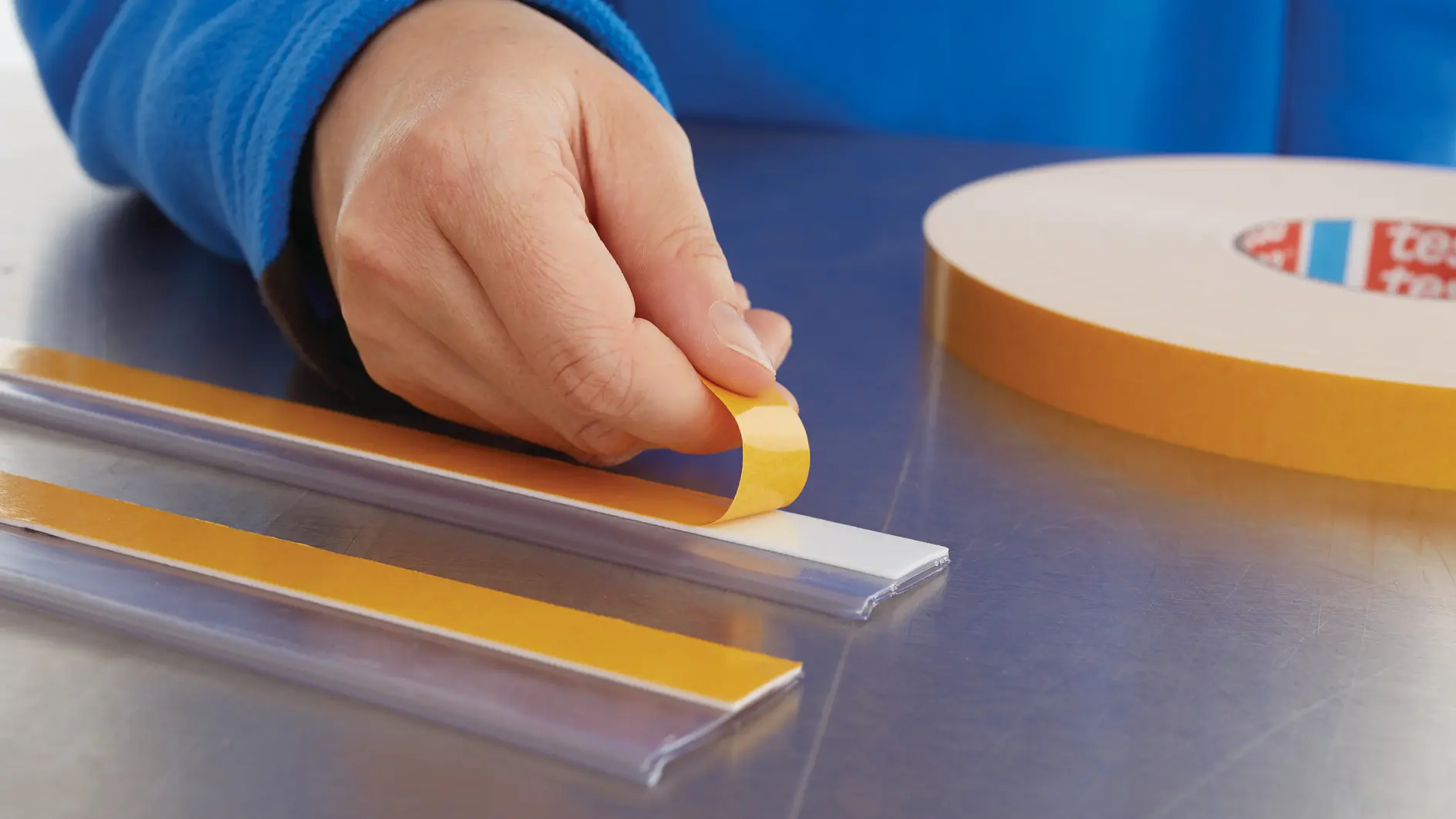mână decojind bandă tesa galbenă dublu adezivă de pe rolă pe o suprafață metalică, cu fâșii de plastic transparente în apropiere (Acest text a fost generat de inteligența artificială)