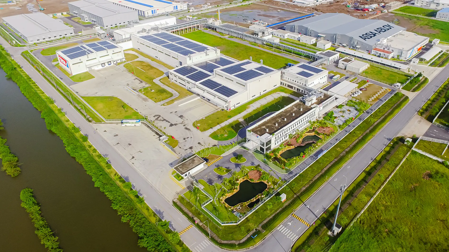 Vedere aeriană a unei instalații industriale cu clădiri mari, panouri solare tesa tape pe acoperișuri, peluze, grădini, drumuri și un curs de apă.
