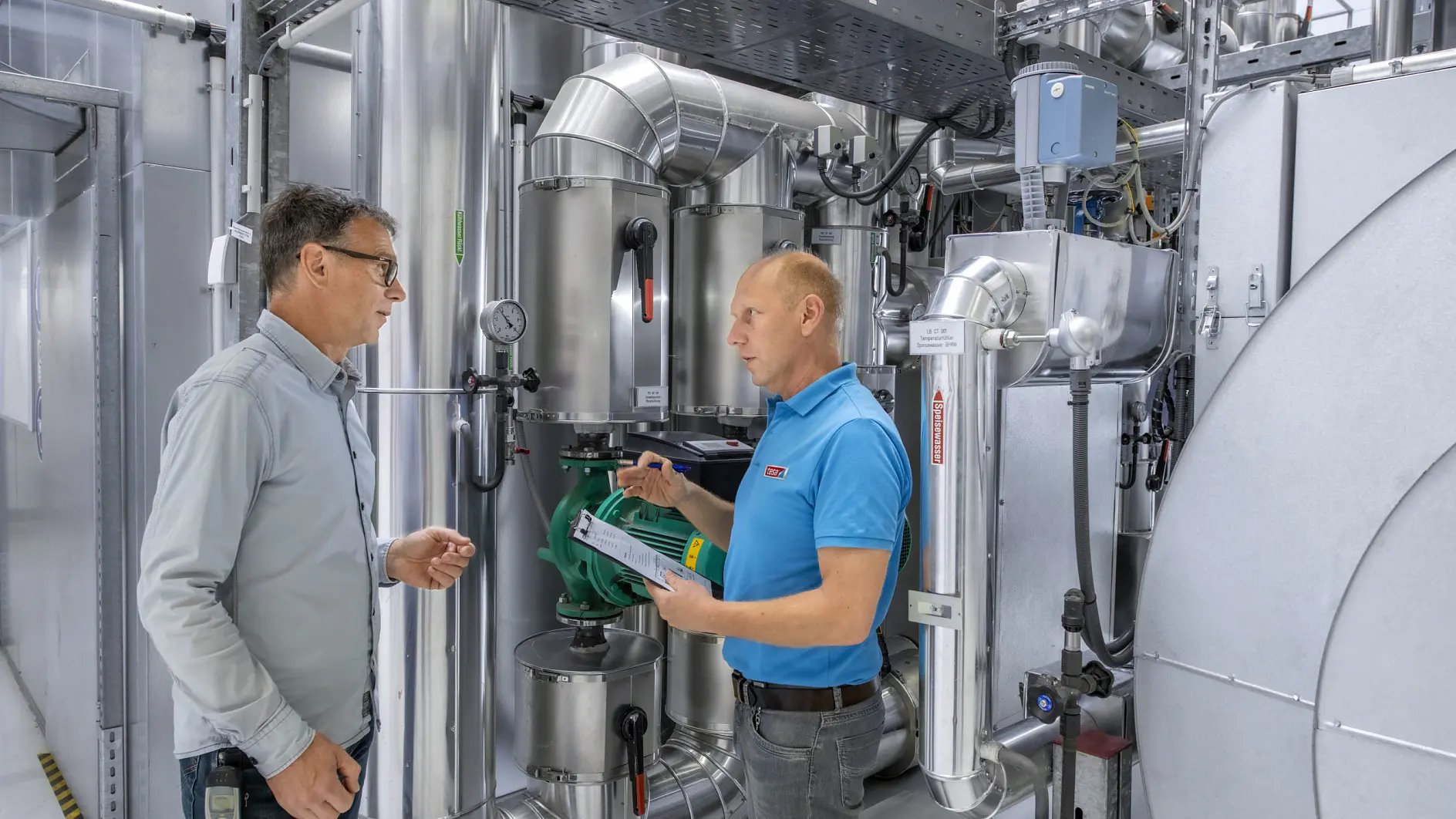 Two men in a technical room discuss operating equipment. One holds a notepad and taps a green valve. They are surrounded by metallic pipes, ducts, and machinery, indicating an industrial or mechanical setting.