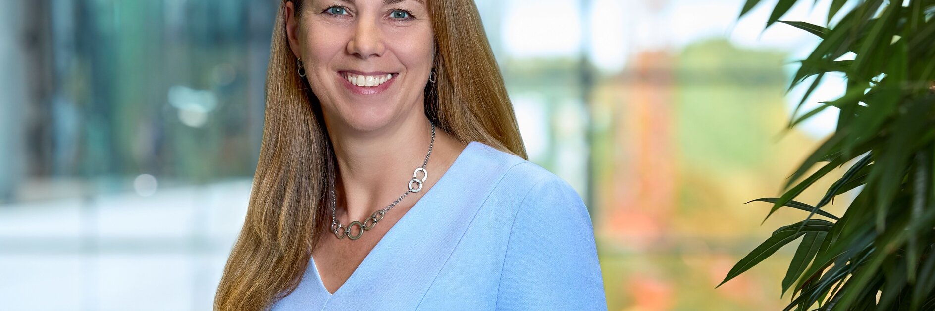A woman with long brown hair stands in a modern hallway lined with large windows. She is wearing a black suit and is leaning on a wooden railing, which has been securely reinforced using tesa tape. The view outside shows buildings and trees. (This text has been generated by AI)