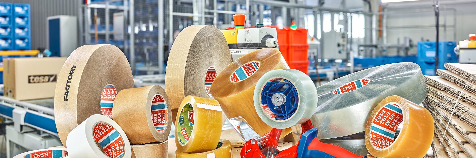 A variety of packing tesa tapes, including clear and brown rolls, are displayed on top of stacked cardboard boxes in an industrial setting. A blue tesa tape dispenser is also present. Shelving and equipment are visible in the background.