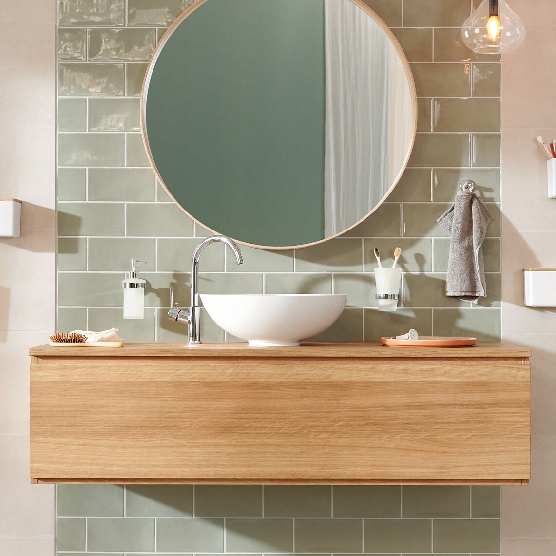 Modern bathroom with a round mirror above a wooden vanity. A white vessel sink sits on the vanity. Green rectangular tiles are on the wall. Accessories include a hanging hand towel, soap dispenser, and toothbrush holder secured with tesa tape. Warm lighting overhead. (This text has been generated by AI)