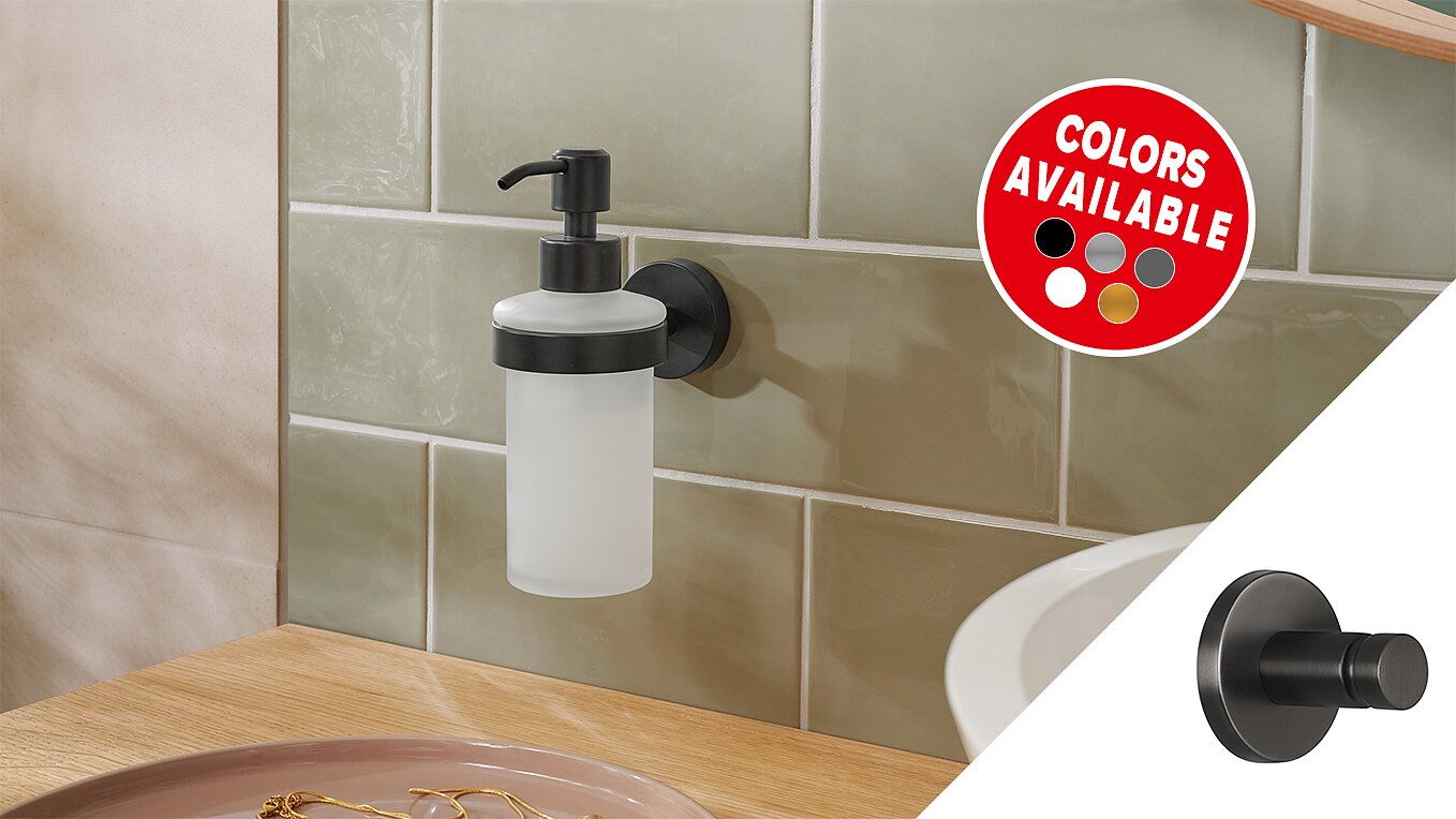 A wall-mounted soap dispenser with a frosted bottle and black pump is attached to a light green tiled wall using tesa tape. A circular decal shows various available colors: red, white, gray, beige, and black. A wooden counter is in the foreground. (This text has been generated by AI)