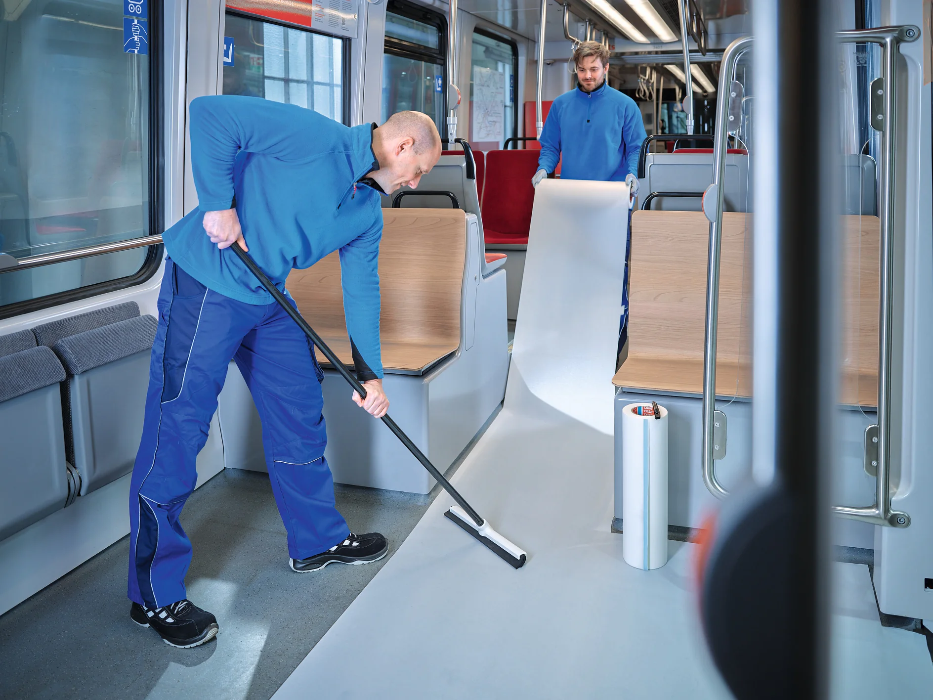 two workers in blue uniforms installing white flooring inside a modern public transport vehicle, one rolling out material while the other smooths it (This text has been generated by AI)