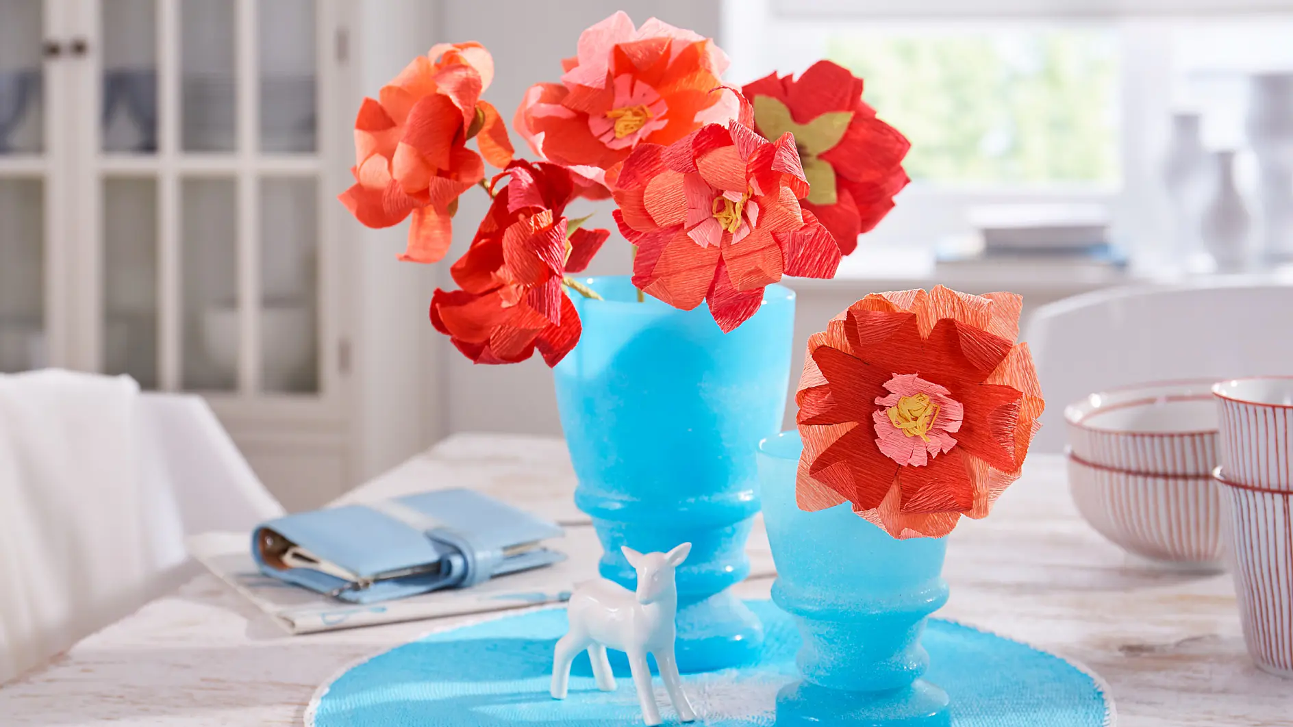 Klarröda och orangea pappersblommor i blå frostade vaser står på ett bord tillsammans med en liten vit hjortfigur. En blå bok och en spiralbunden anteckningsbok ligger bredvid till höger. I bakgrunden syns hyllor med porslin, prydligt säkrade med tesa-tejp. (This text has been generated by AI)
