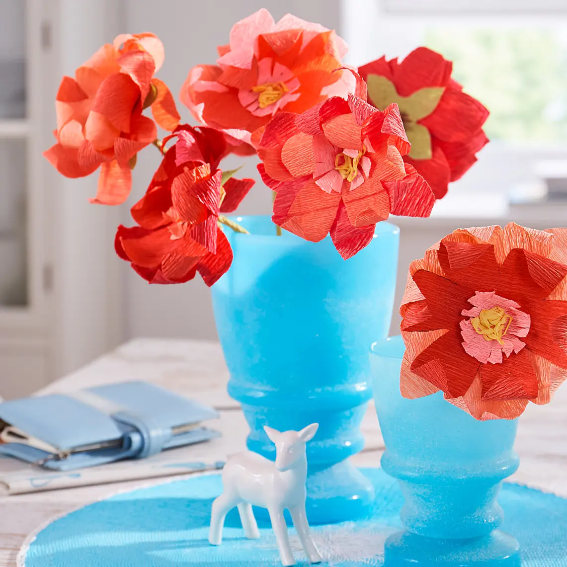 Klarröda och orangea pappersblommor i blå frostade vaser står på ett bord tillsammans med en liten vit hjortfigur. En blå bok och en spiralbunden anteckningsbok ligger bredvid till höger. I bakgrunden syns hyllor med porslin, prydligt säkrade med tesa-tejp. (This text has been generated by AI)