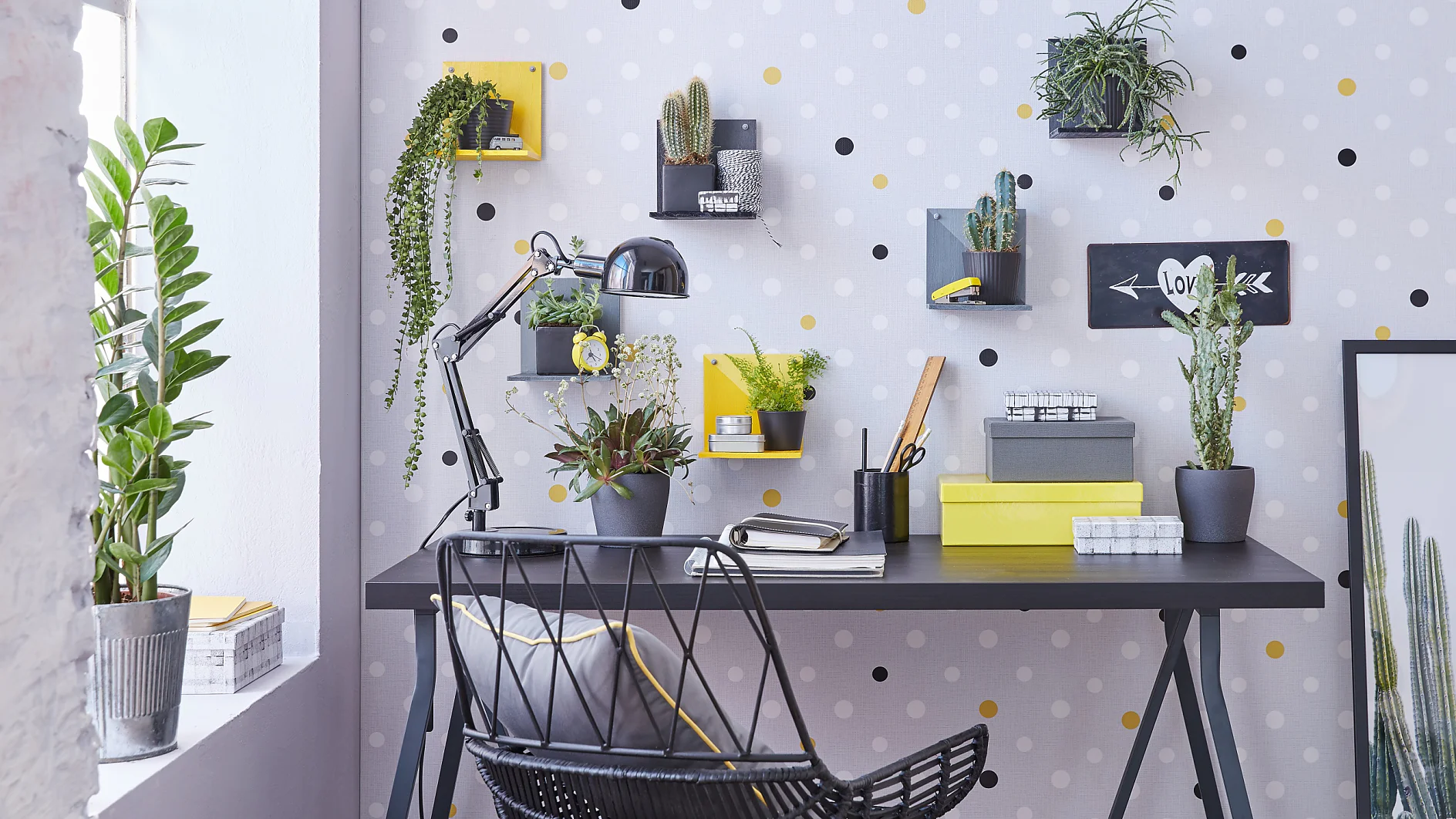 A modern desk setup with a black tabletop, two notebooks, a desk lamp, and several potted plants. The wall has a polka dot pattern with small shelves holding plants and decor. A wicker chair with a gray cushion is placed at the desk, seamlessly assembled using tesa tape for durability and style. (This text has been generated by AI)