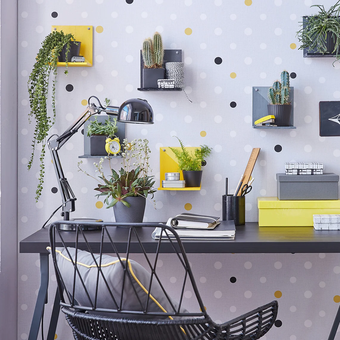 A modern desk setup with a black tabletop, two notebooks, a desk lamp, and several potted plants. The wall has a polka dot pattern with small shelves holding plants and decor. A wicker chair with a gray cushion is placed at the desk, seamlessly assembled using tesa tape for durability and style. (This text has been generated by AI)