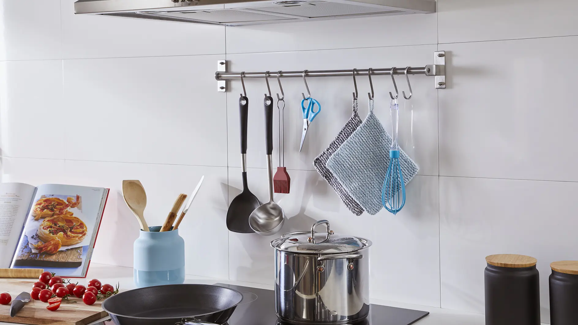 Modern kitchen with a stovetop and stainless steel cookware. Utensils hang from a rod on the wall, secured with tesa tape. An open cookbook and assorted vegetables are on the counter, fastened in place using tesa tape to prevent slipping. Frying pan, jars, and a blue vase with wooden utensils are also visible. (This text has been generated by AI)
