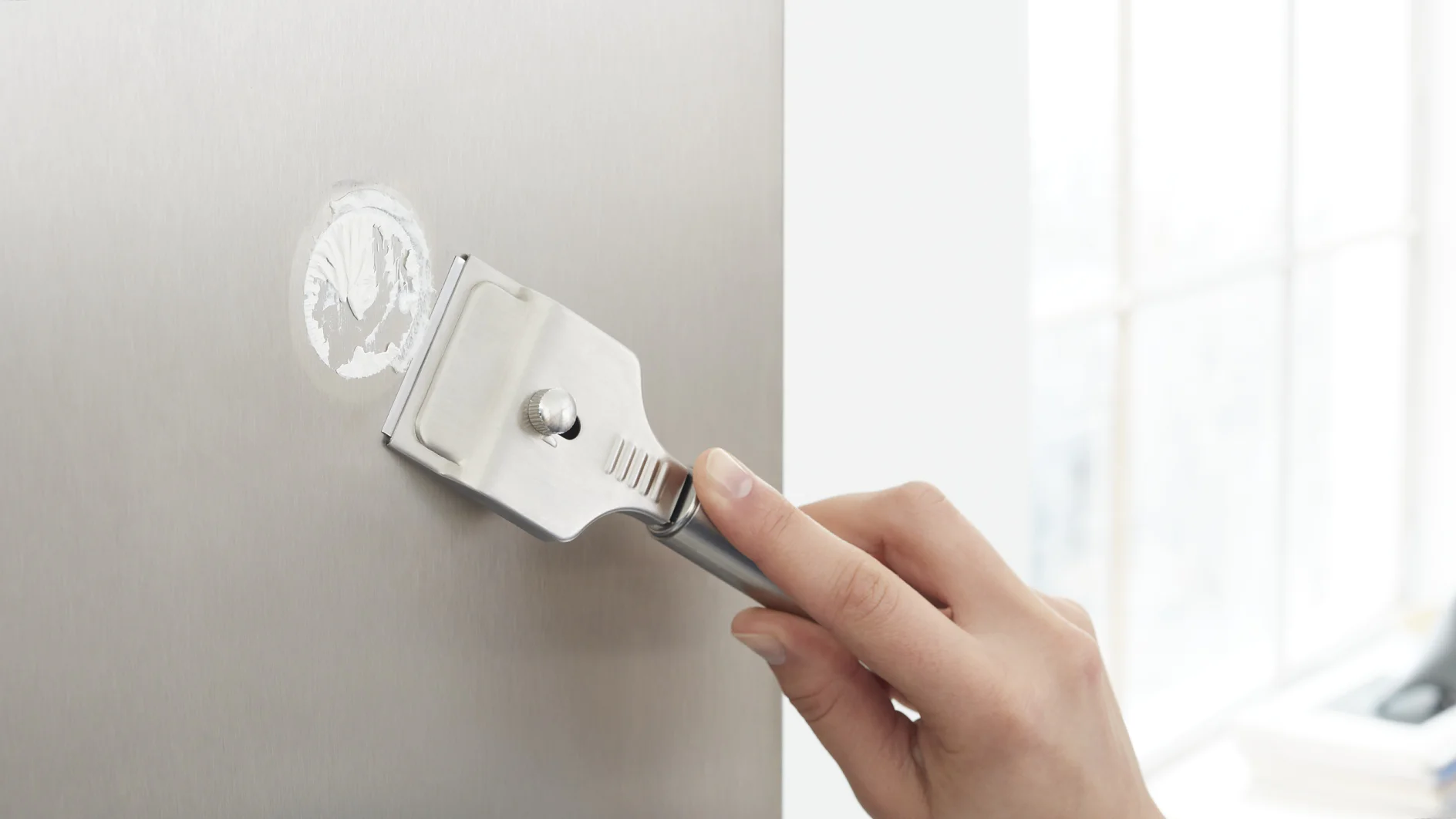A hand is using a metal scraper to remove a circular sticker from a smooth metal surface where tesa tape was previously applied. The background is softly lit, possibly by natural light coming through a window. (This text has been generated by AI)