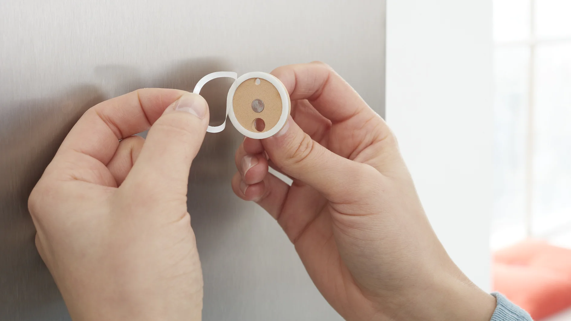 Two hands are applying a round adhesive sticker with two cut-out holes onto a metallic surface using tesa tape. The sticker has a white edge and a brown center. One hand is peeling off the backing of the sticker. (This text has been generated by AI)