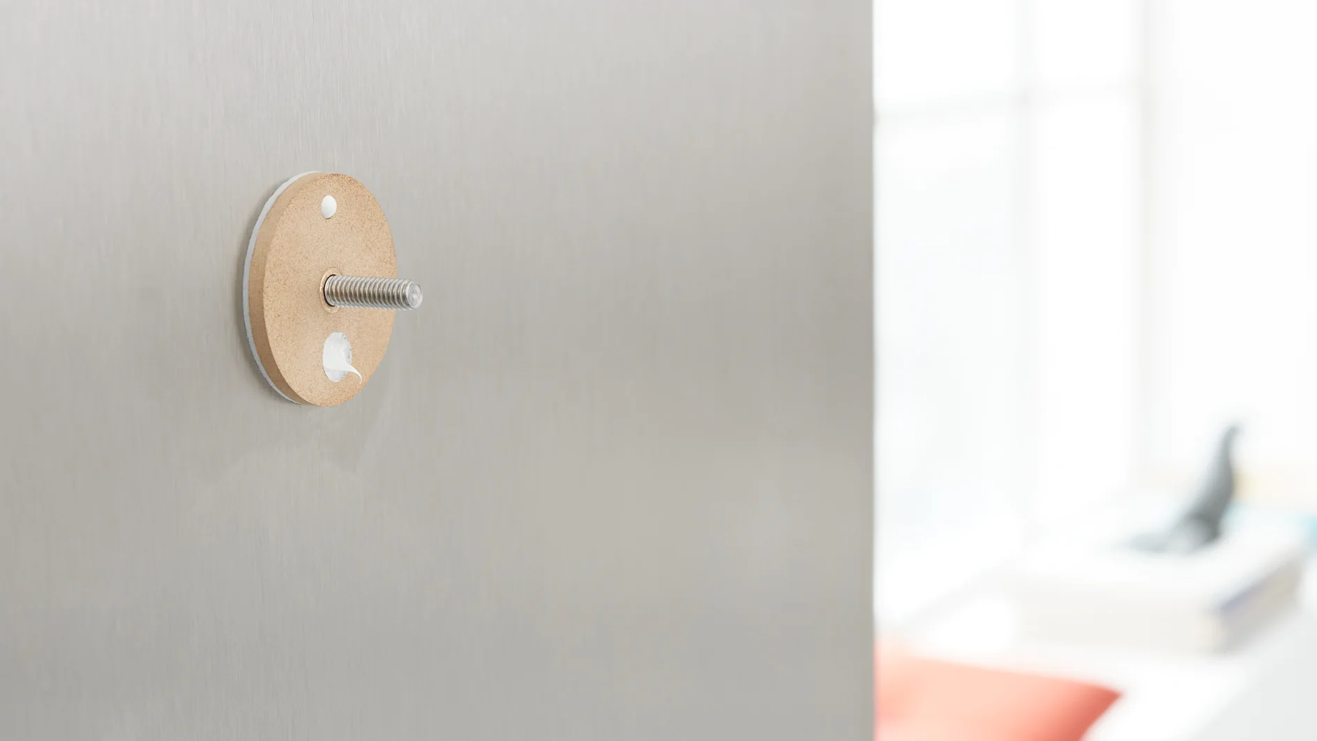 Close-up of a metal surface with a protruding bolt. The bolt passes through a circular piece of cardboard secured with tesa tape. The background is blurred, showing part of a window and some indistinct objects. (This text has been generated by AI)