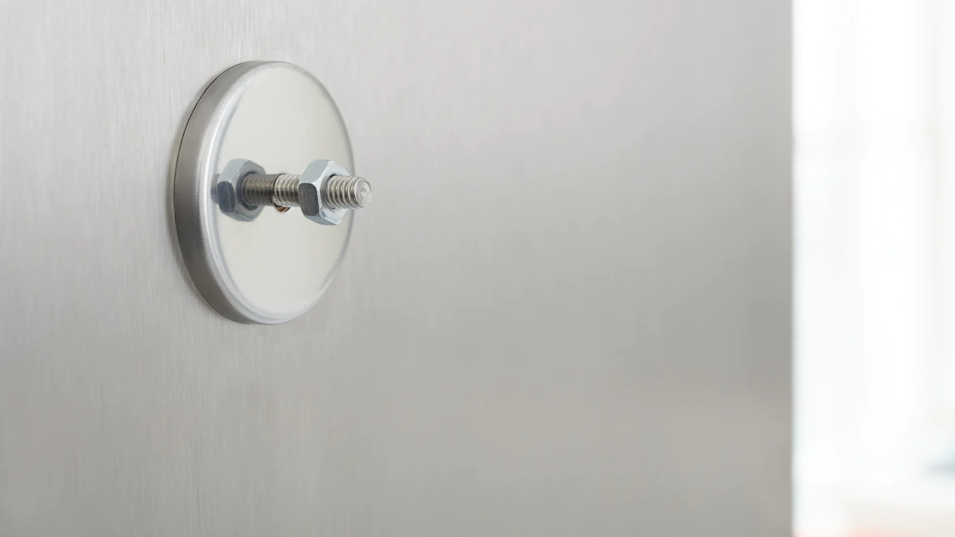 A close-up image of a silver bolt and nut secured into a circular metal plate, set against a smooth, light gray metallic surface. The background appears softly blurred, emphasizing the hardware components with tesa tape used to achieve secure fastening. (This text has been generated by AI)