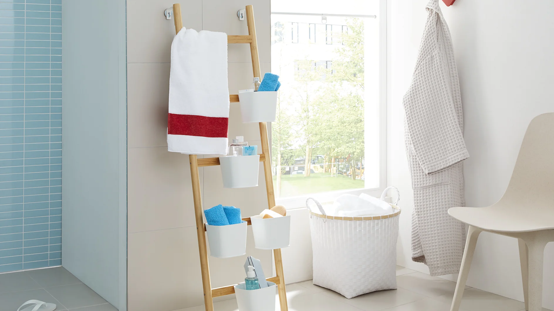 Bathroom with a light, airy atmosphere. Towels and toiletries are organized on a wooden ladder shelf. A white laundry basket and robe are nearby. A window provides natural light, and a chair and slippers complete the cozy setting. (This text has been generated by AI)