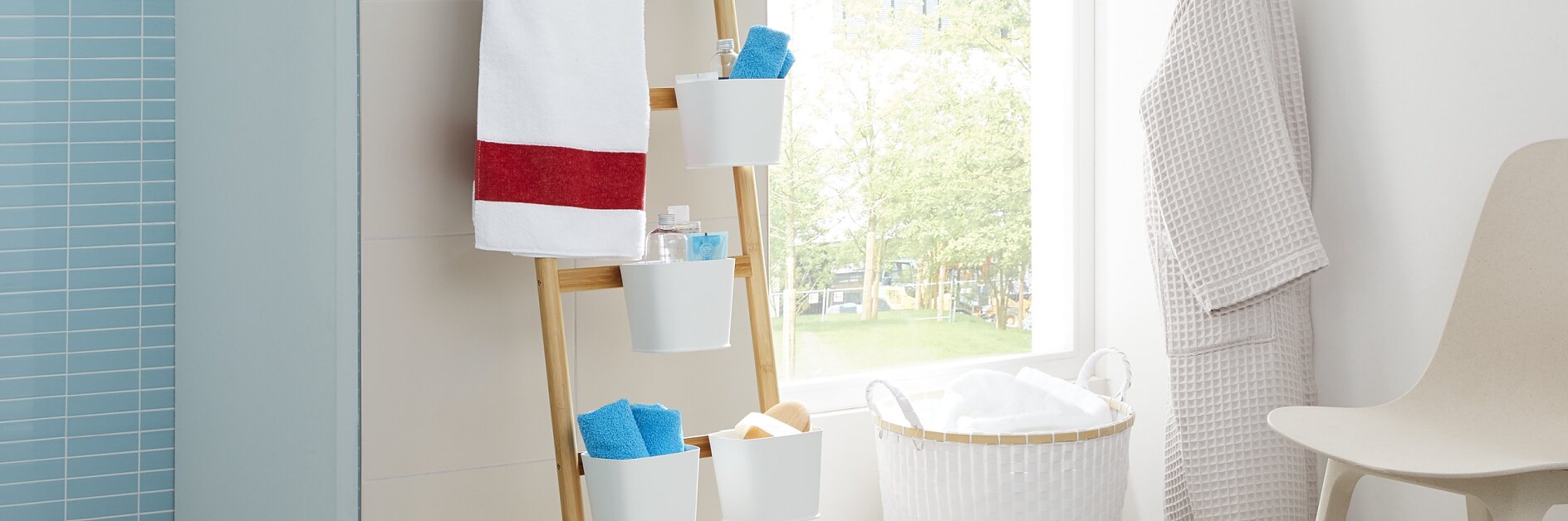 Bathroom with a light, airy atmosphere. Towels and toiletries are organized on a wooden ladder shelf. A white laundry basket and robe are nearby. A window provides natural light, and a chair and slippers complete the cozy setting. (This text has been generated by AI)