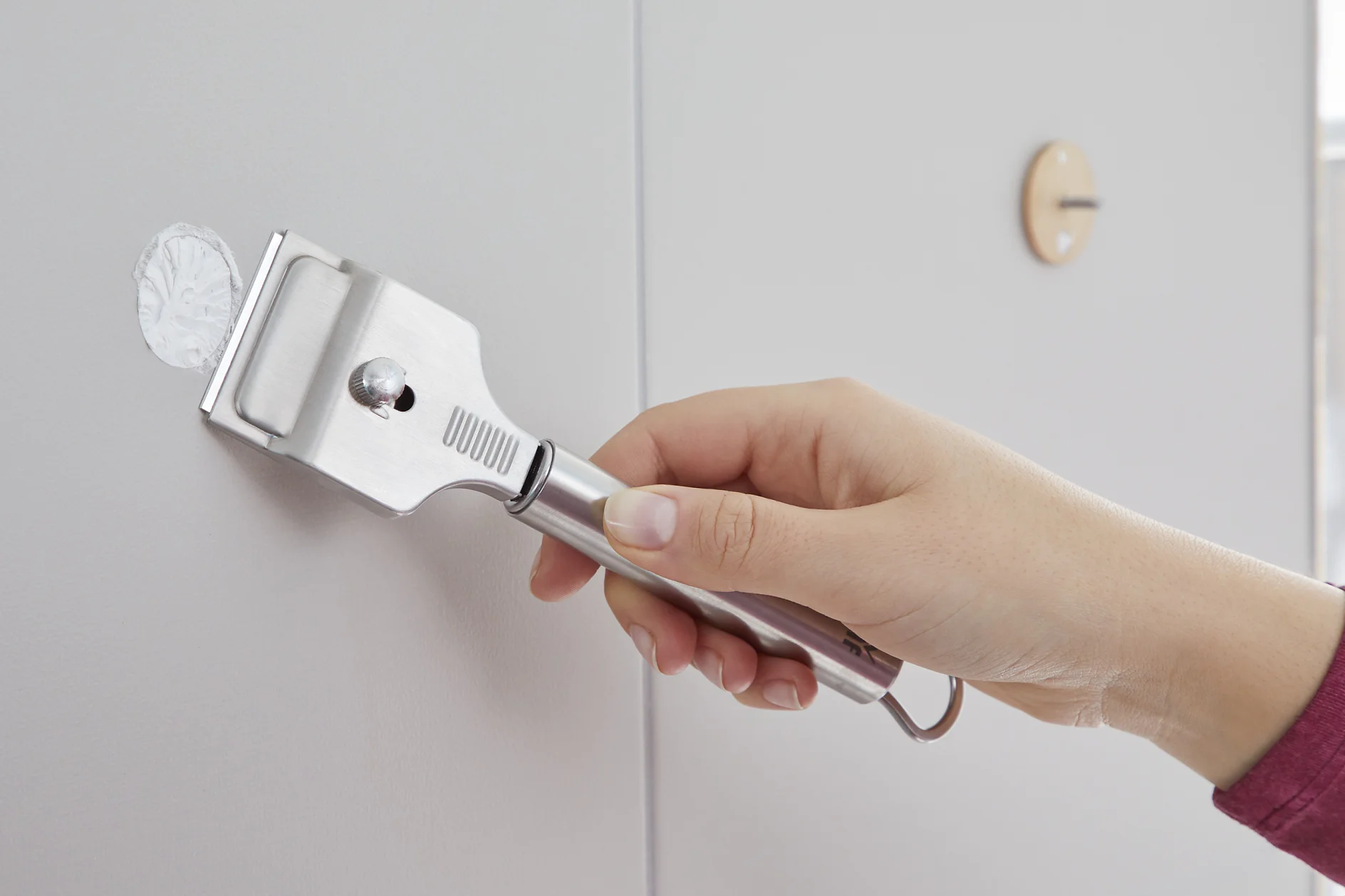 A hand holding a metal wall scraper is applying a layer of joint compound to a small circular area on a wall. There is a round protective sticky tab made of tesa tape on the wall near the applied area. The wall is light gray. (This text has been generated by AI)