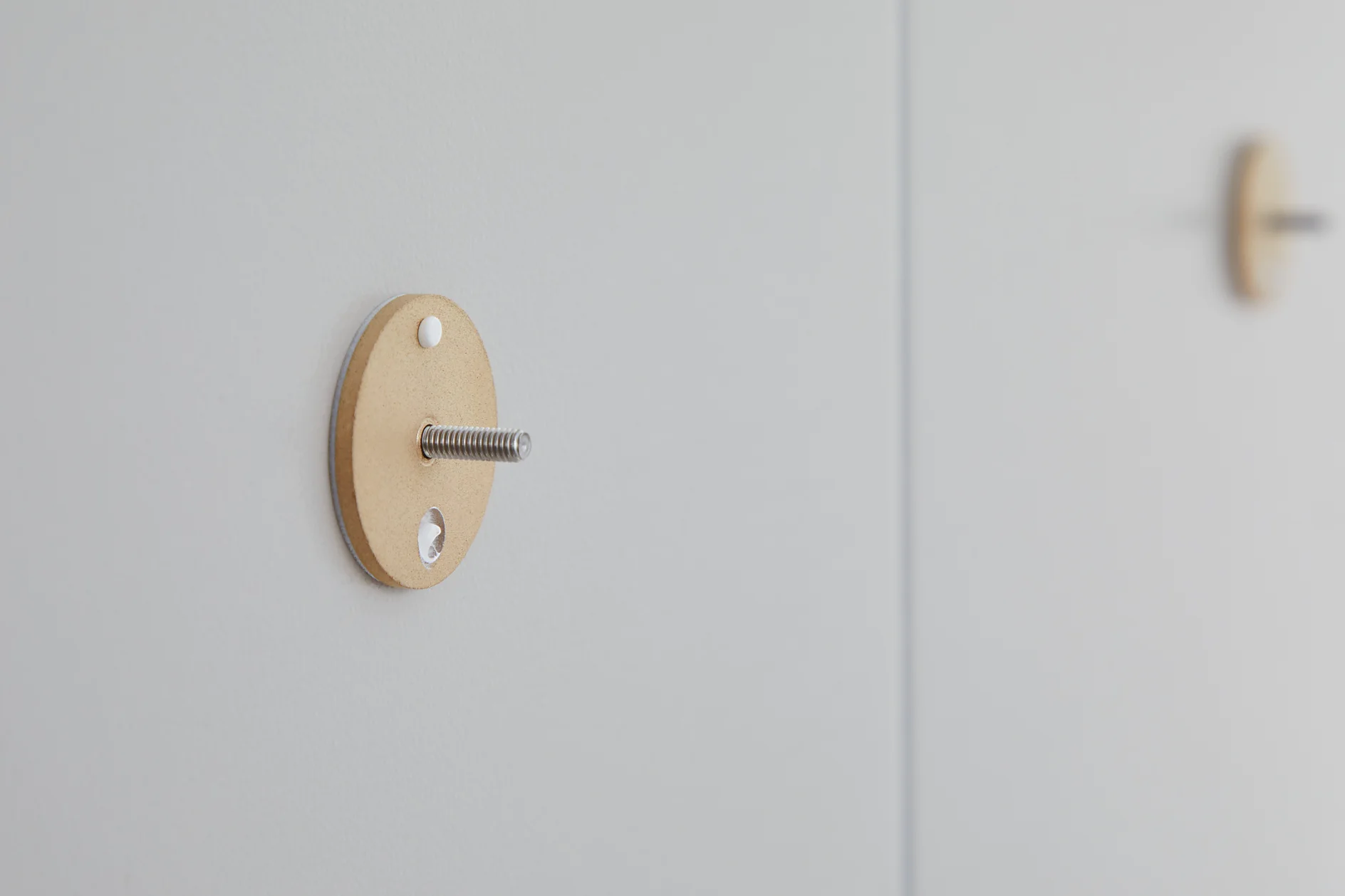 A small circular wooden disc with a screw protruding from its center is mounted on a light gray wall using tesa tape. Another similar fixture is slightly blurred in the background, also secured with tesa tape. (This text has been generated by AI)