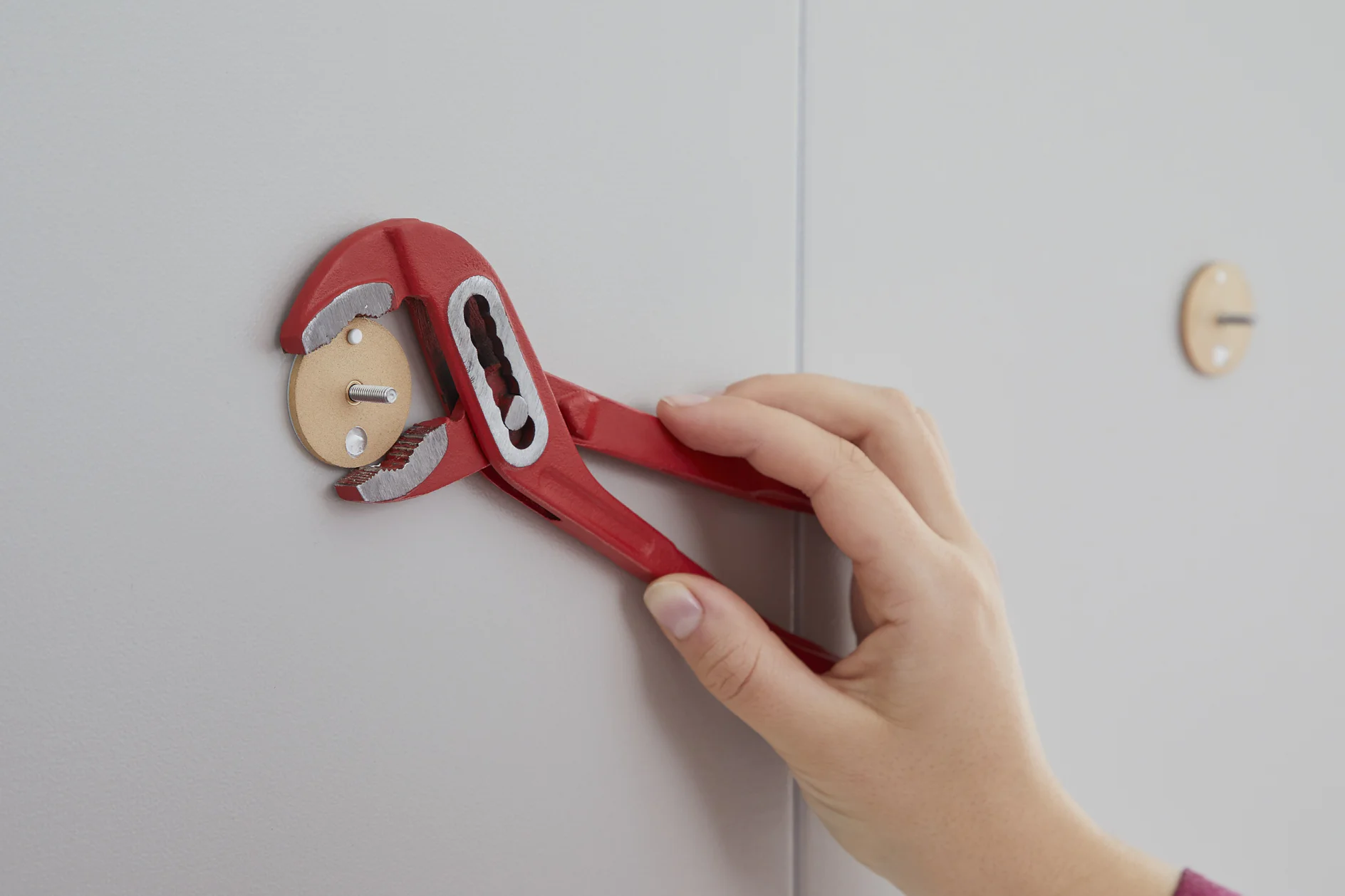A hand uses a red adjustable wrench to tighten or loosen a small assembly mounted on a light gray wall. The assembly has a circular wooden backing and two small pegs secured with tesa tape. Another similar assembly, also held in place by tesa tape, is visible in the background. (This text has been generated by AI)