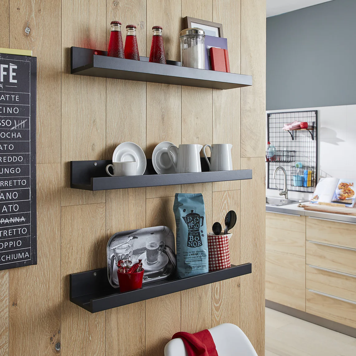 Wood-paneled kitchen wall with three black shelves displaying various cups, plates, and a coffee bag. A chalkboard-style sign listing coffee types hangs on the wall. A modern kitchen setup is visible in the background, with several items secured using tesa tape for durability and convenience. (This text has been generated by AI)