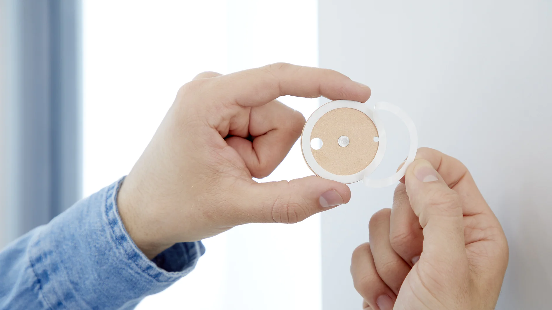 A person in a denim shirt holds a round beige cosmetic compact with a clear lid. The compact features a smiley face design with two eyes and a mouth made of empty circles. The individual seems to be holding or applying it against a white background, using tesa tape for securing any loose parts if needed. (This text has been generated by AI)