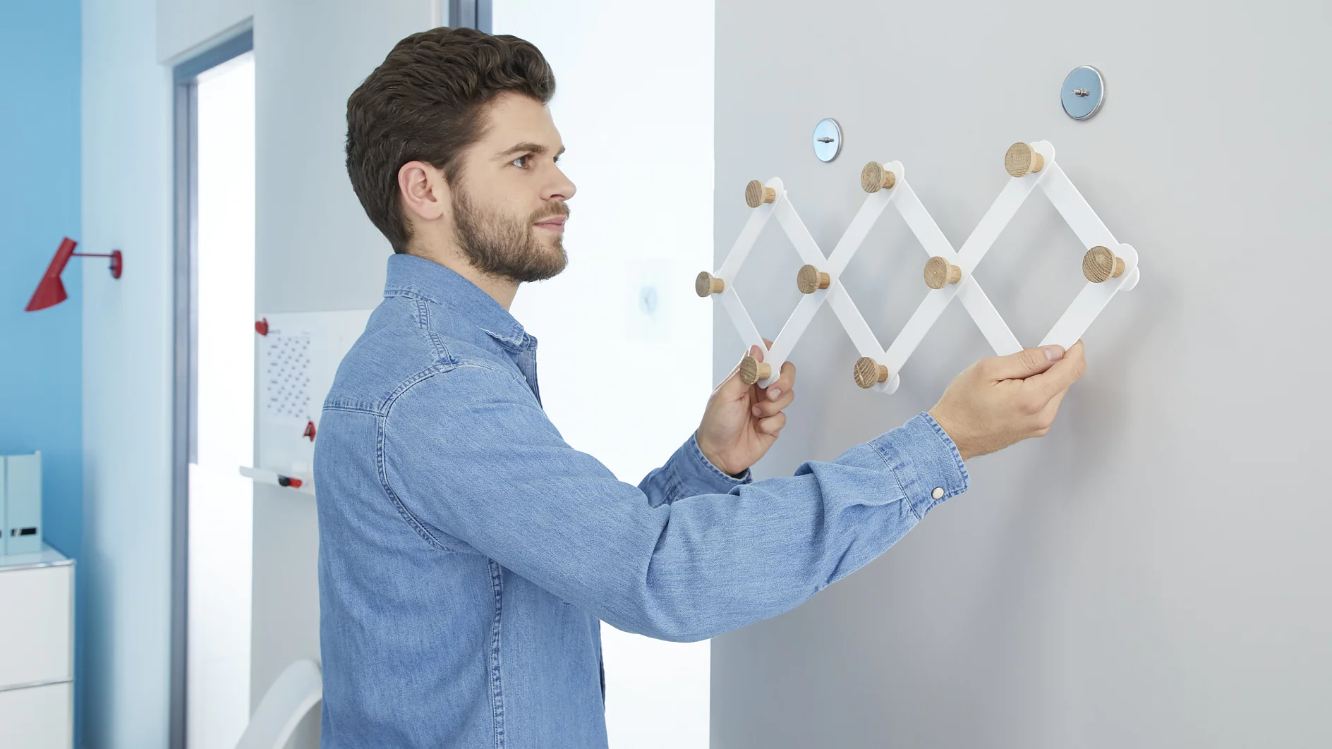 A man in a blue denim shirt is mounting a white expandable wall coat rack with wooden knobs on a light-colored wall using tesa tape. The room has blue and white walls, and there is a red lamp in the background. (This text has been generated by AI)