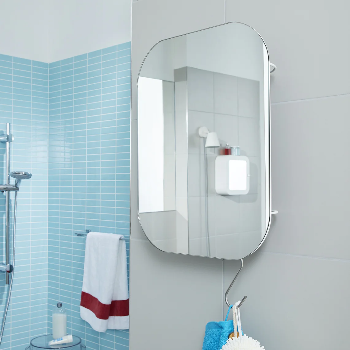 A modern bathroom features light blue tiled walls and a glass shower enclosure. A rounded rectangular mirror is mounted on a gray wall, secured in place with tesa tape. A towel hangs below the mirror. Sunlight filters in through a window on the right. (This text has been generated by AI)