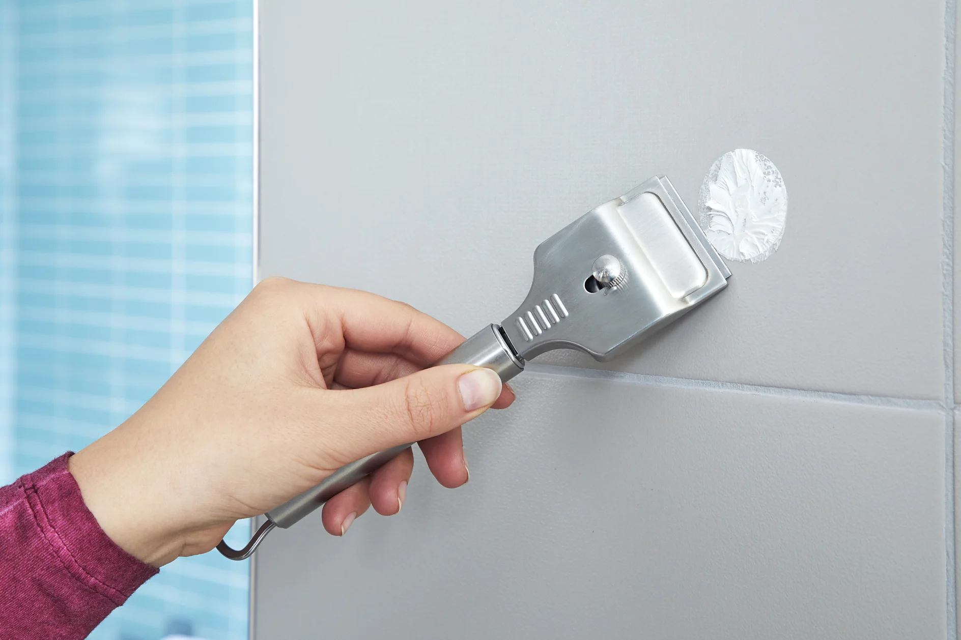 A hand holds a metal scraper in front of a tiled wall, removing tesa tape adhesive from the surface. The person is wearing a maroon sleeve. The tiles are gray with white grout, and there is a blurry background of blue patterned tiles. (This text has been generated by AI)