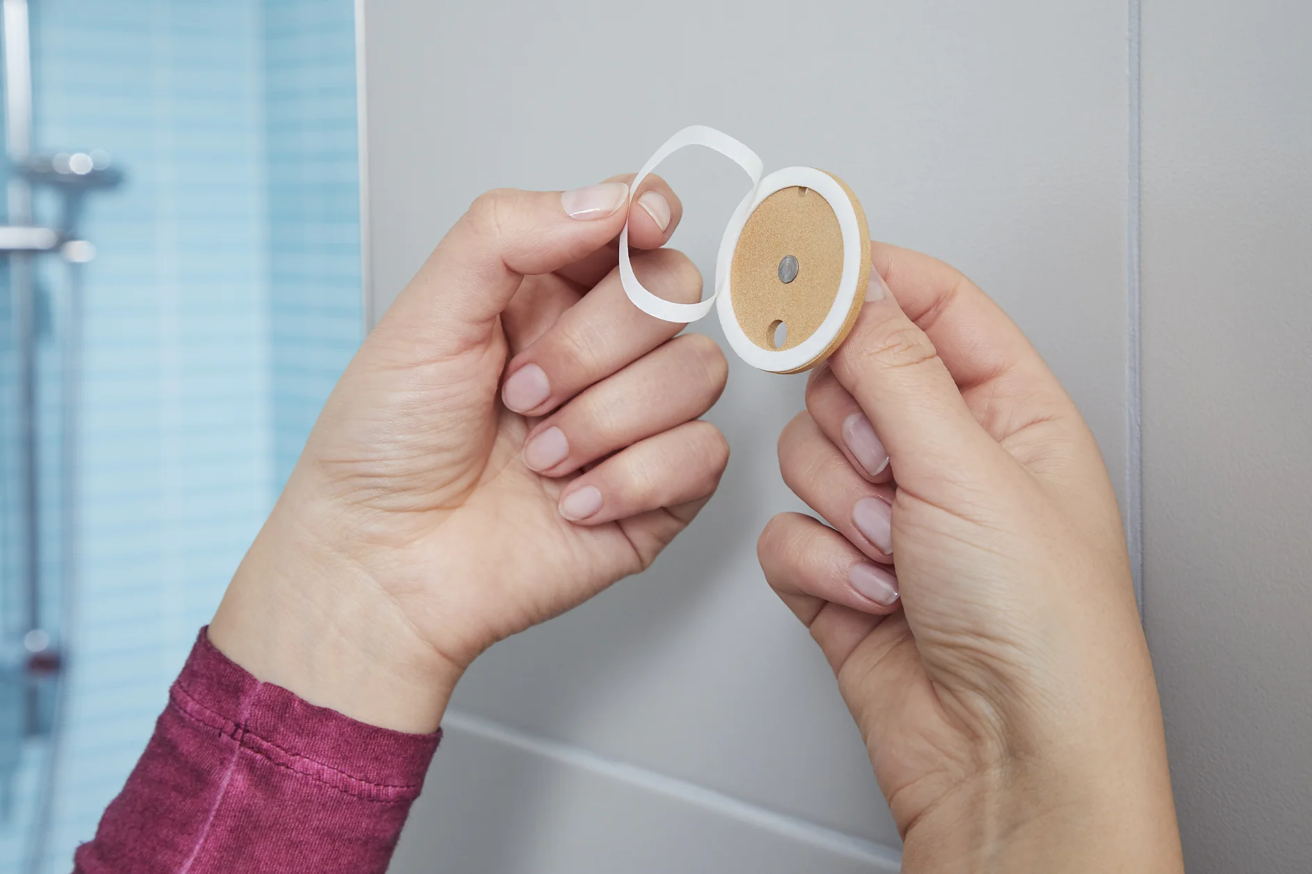Close-up of hands peeling the backing from a tesa tape adhesive disc with a central hole. The background features a tiled wall with a blue hue. The person is wearing a long-sleeved pinkish-red top. (This text has been generated by AI)