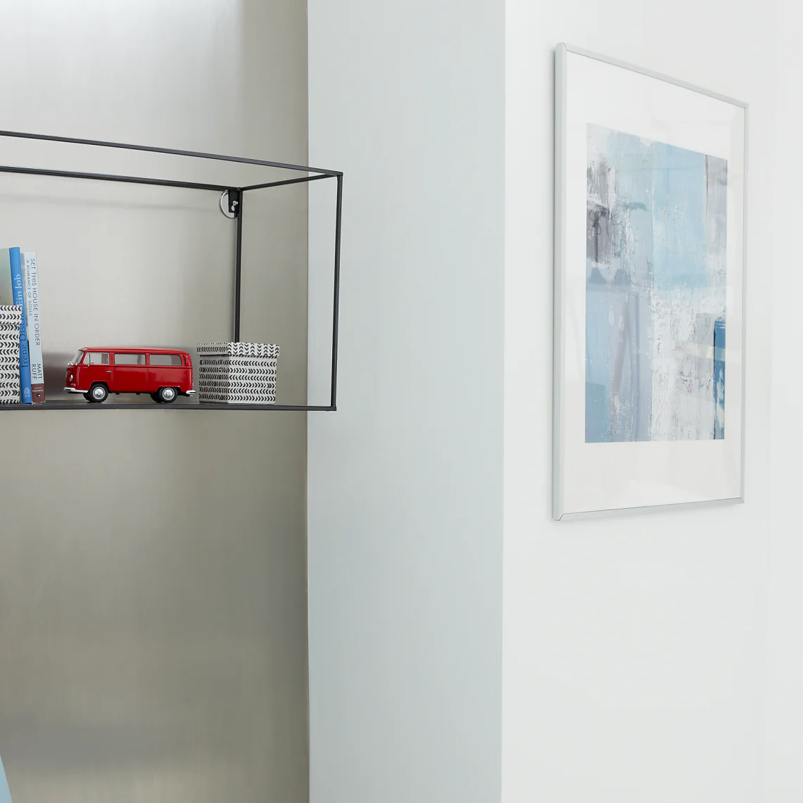 A minimalist room features a wall-mounted shelf secured with tesa tape, holding books, a small red van model, and a decorative box. A framed abstract art piece hangs on the wall with the help of tesa tape, and a modern black floor lamp is nearby. There is a blue chair partially visible. (This text has been generated by AI)