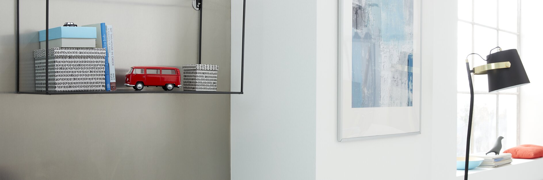 A minimalist room features a wall-mounted shelf secured with tesa tape, holding books, a small red van model, and a decorative box. A framed abstract art piece hangs on the wall with the help of tesa tape, and a modern black floor lamp is nearby. There is a blue chair partially visible. (This text has been generated by AI)