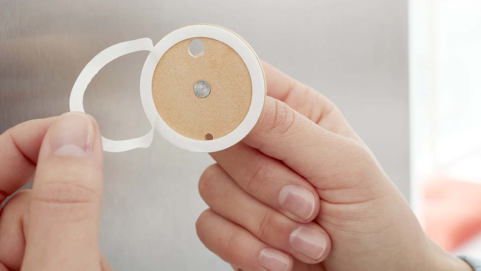 A hand holding a round adhesive-backed device with a central sensor and two cut-out holes near the top. The device is positioned against a blurred gray background, secured in place using tesa tape. (This text has been generated by AI)