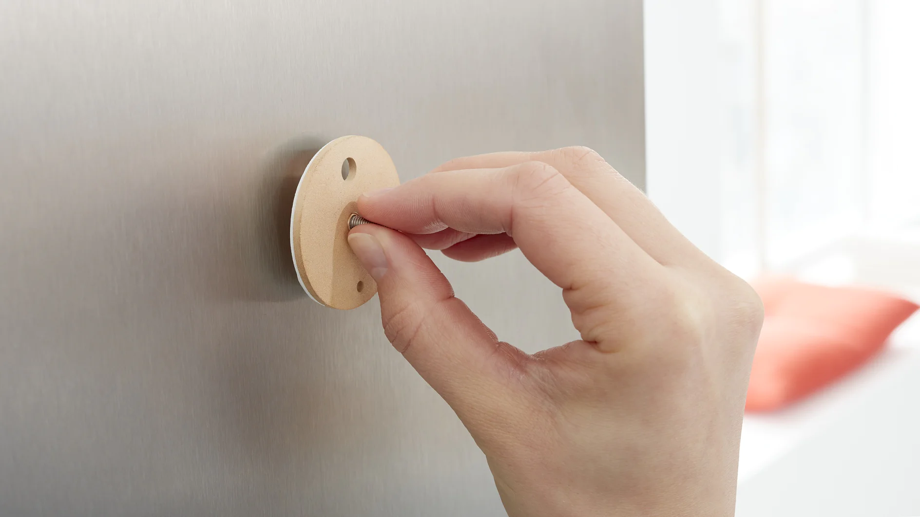A hand is turning a beige, circular dial attached to a smooth, metallic surface. The dial has three small holes around its edge. The background is softly blurred, with a hint of red in the distance. (This text has been generated by AI)