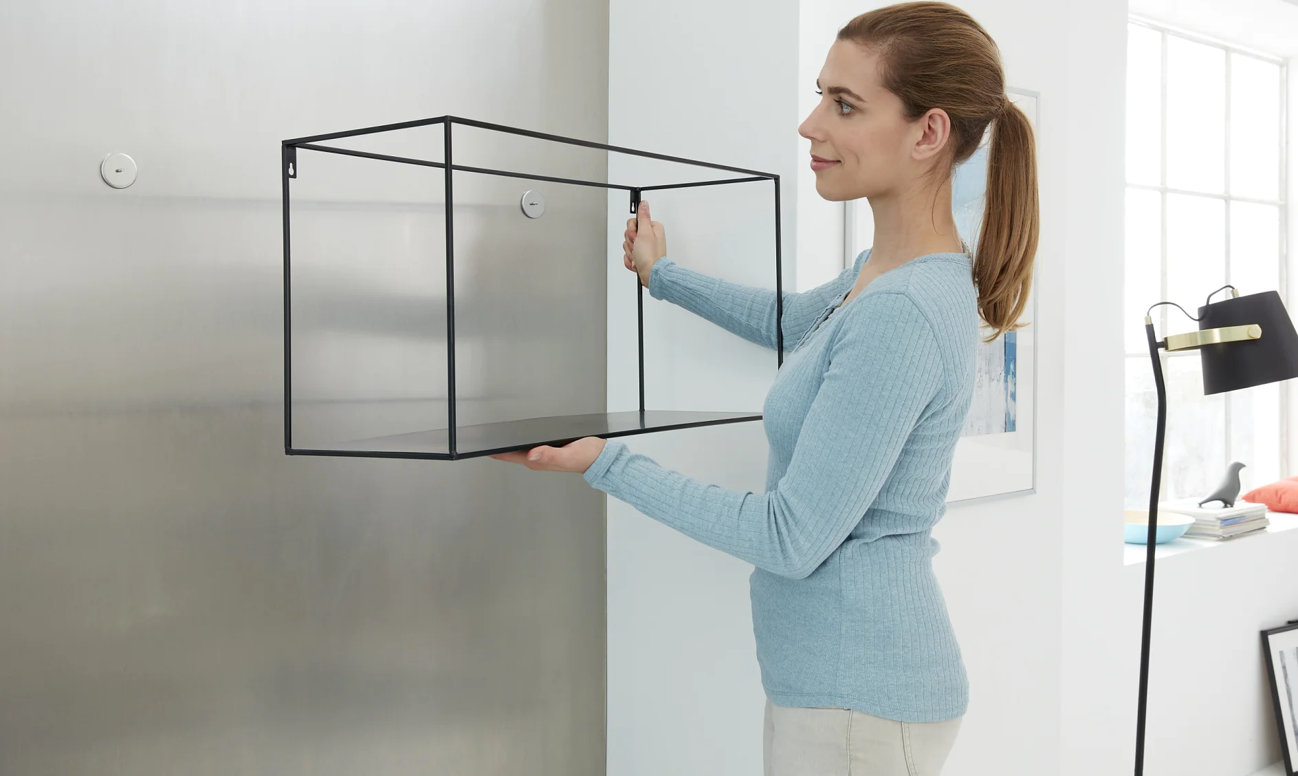 A woman in a blue sweater is holding a large, transparent glass and metal frame structure, positioning it against a white and metallic wall. She is standing in a modern, bright room with minimalistic decor, using tesa tape to secure the structure in place. (This text has been generated by AI)