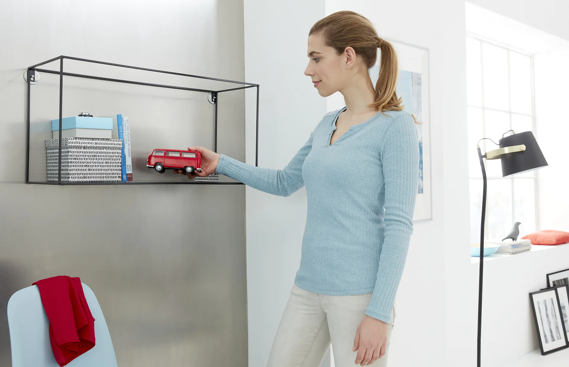 A woman in a light blue sweater places a red toy van on a black metal shelf. The shelf holds books and a box. A red cloth is draped over a nearby chair, secured with tesa tape. A black floor lamp is visible by the window in the background, with tesa tape adding decorative accents. (This text has been generated by AI)